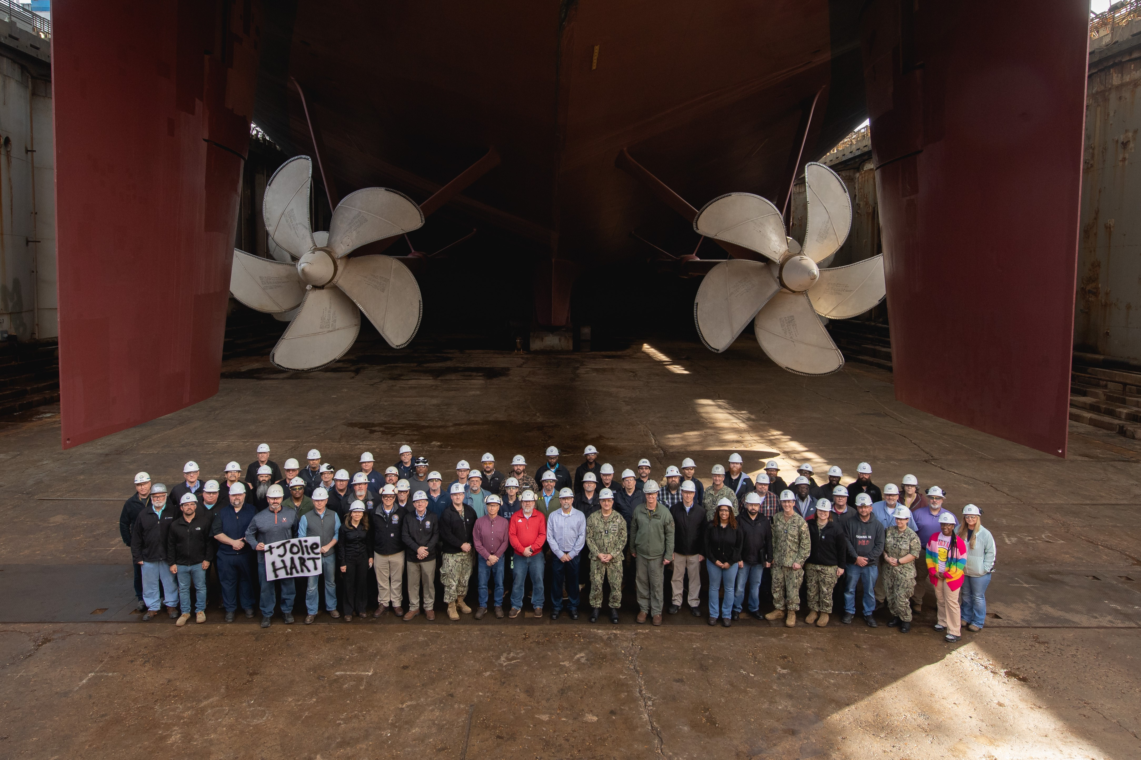 image of a large group of individuals with hardhats on. They are standing in front of a PEO carrier in Newport News, Virginia. Two large propellers are featured. Two of the individuals hold a sign that reads